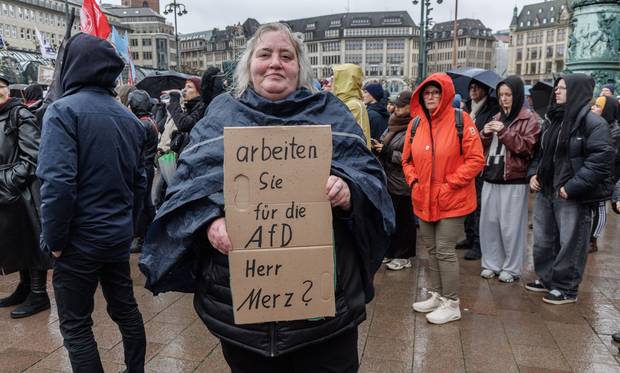 Eine Demonstrantin in Hamburg stellt auf einem Pappschild eine Frage an Bundeskanzler Friedrich Merz (CDU).